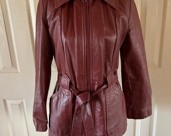 Può includere: Una giacca di pelle bordeaux con zip frontale e cintura. La giacca ha un colletto e due tasche.