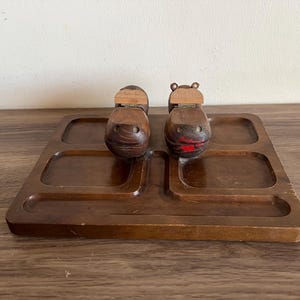 May include: A wooden serving tray with four compartments, holding two carved wooden hippopotamus salt and pepper shakers. The hippos have red painted lips. The tray and shakers are a dark brown colour.