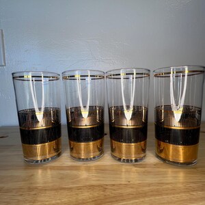 May include: Four vintage highball glasses. Each glass is clear with a gold rim and gold and black bands around the middle and base. The glasses are arranged on a wooden surface.