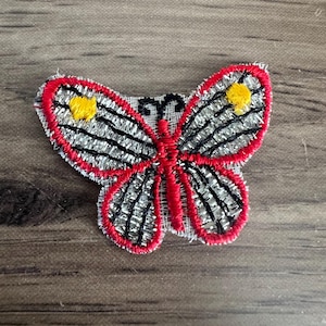 May include: A red and silver butterfly applique with yellow accents. The butterfly is embroidered with a detailed design and has a delicate, sheer fabric.