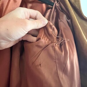 May include: Close-up of the inside of a brown suede jacket, showing the lining. The lining is a light brown color and is made of a smooth fabric. There is a small hole in the lining.