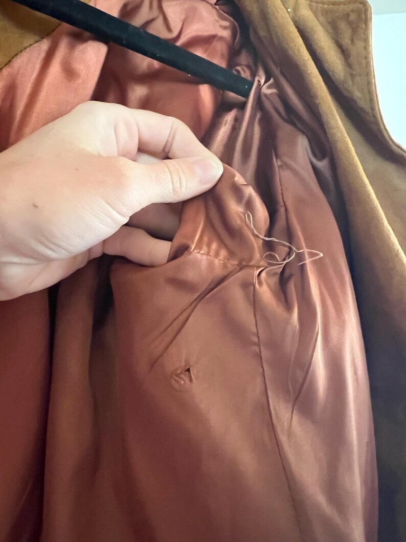 May include: Close-up of the inside of a brown suede jacket, showing the lining. The lining is a light brown color and is made of a smooth fabric. There is a small hole in the lining.
