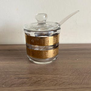 May include: A clear glass jar with a gold geometric pattern and a matching lid. The jar has a small spoon attached to the lid.