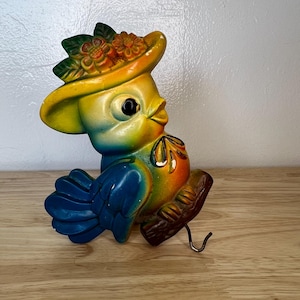May include: A colorful ceramic bird figurine wearing a yellow hat adorned with orange flowers and green leaves. The bird has blue, yellow, and orange hues, perched on a brown branch with a metal hook. A whimsical decorative item.