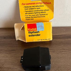 May include: A vintage Kodak flipflash extender kit. The yellow and white box reads "Kodak flipflash extender" and includes text about reducing red reflections. A black plastic flash unit sits below the box.