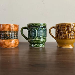 May include: Three ceramic mugs with unique designs. The mug on the left is orange with a geometric pattern, the middle mug is green with a rope design, and the right mug is brown with a brick pattern. Each mug has a curved handle.
