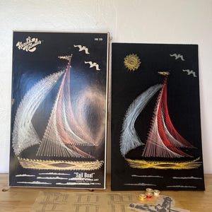 May include: A vintage "Sail Boat" dimensional string art kit and finished piece. The artwork features a sailboat design with white, red, and gold thread on a black background. The kit includes a box, instructions, and spools of thread.