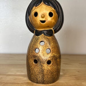 May include: A vintage ceramic doll-shaped candle holder. The doll has a gold-colored face, a black bow, and black hair. The body is gold with three circular openings. The candle holder is on a wooden surface.
