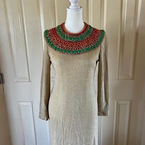 Puede incluir: Un vestido largo de manga larga dorado brillante con un gran collar de cuentas verdes y naranjas.