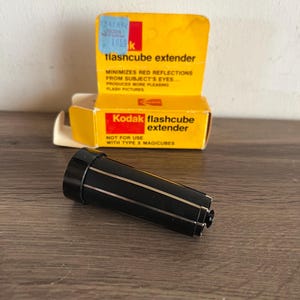 May include: A black flashcube extender with gold stripes, next to a yellow Kodak box. The box reads "flashcube extender" and "MINIMIZES RED REFLECTIONS FROM SUBJECT'S EYES... PRODUCES MORE PLEASING FLASH PICTURES."