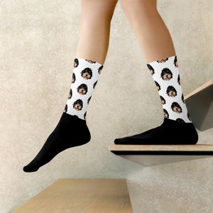 May include: A pair of white socks with a black and brown dog face pattern. The socks are worn on a person's feet, with the person's legs visible.