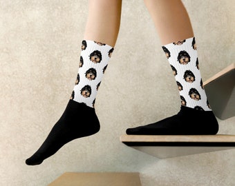 Puede incluir: Un par de calcetines blancos con un patrón de cara de perro negro y marrón. Los calcetines se llevan puestos en los pies de una persona, con las piernas de la persona visibles.