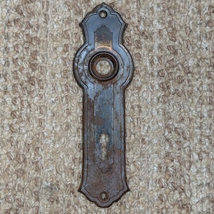 Puede incluir: Una placa de puerta de metal oscuro y desgastado con una abertura circular central para un pomo y un corte de cerradura en la parte inferior. La placa rectangular alargada tiene extremos decorativos redondeados con orificios para tornillos.
