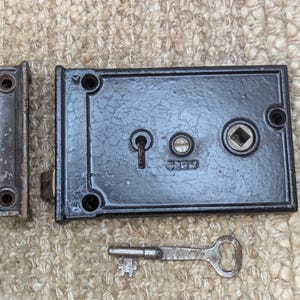 May include: Black cast iron door lock with a keyhole and a square key slot. The lock has a black metal strike plate and comes with a silver key.