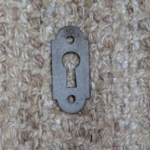 May include: A dark gray metal keyhole escutcheon plate with a rectangular shape featuring rounded top and bottom edges. The plate has a central keyhole cutout and two circular screw holes, one positioned above and one below the keyhole opening.