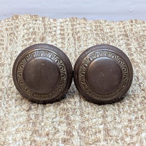 May include: Two antique-style, round, metal door knobs with a decorative, raised, circular design around the edge. The knobs are a dark brown color.