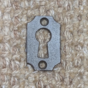 Op de afbeelding: Een kleine, rechthoekige metalen escutcheonplaat in antieke stijl met een sleutelgatvorm in het midden. De plaat heeft twee ronde gaten, één boven en één onder het sleutelgat. Het metaal heeft een getextureerd, verouderd uiterlijk.