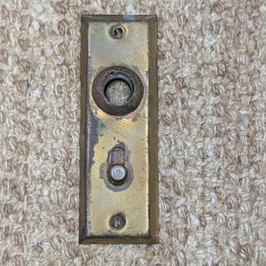 Könnte beinhalten: Eine rechteckige Türplatte aus Messing mit einer zentralen kreisförmigen Öffnung und einem kleinen verschiebbaren Metallriegel darunter. Die Platte hat oben und unten jeweils ein Schraubloch und eine verwitterte, angelaufene Oberfläche.