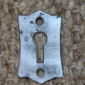 Op de afbeelding: Een zilveren sleutelgatbedekking met een decoratieve, licht puntige, rechthoekige vorm. De afdekking heeft twee kleine gaten aan weerszijden van het sleutelgat.