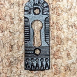 Könnte beinhalten: Schwarzer Schlüsselschlitzabdeckung aus Gusseisen mit dekorativem Design. Die Abdeckung hat ein Schlüsselloch in der Mitte und einen gewellten unteren Rand.