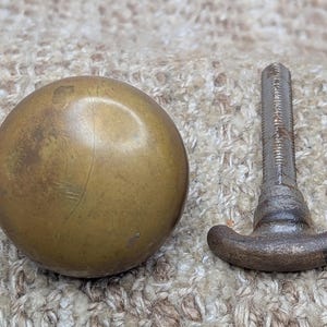 May include: A vintage brass knob with a round, aged appearance, next to a matching metal screw. The knob is a warm, golden color with signs of wear. The screw has a T-shaped handle.