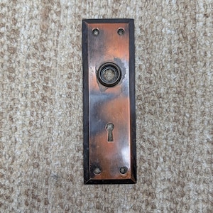 Könnte beinhalten: Eine rechteckige Türschildplatte aus Metall mit kupferfarbenem Finish und dunklen Kanten. Die Platte verfügt über eine zentrale kreisförmige Öffnung für einen Knauf, ein Schlüsselloch darunter und vier kleine Schraublöcher an den Ecken.