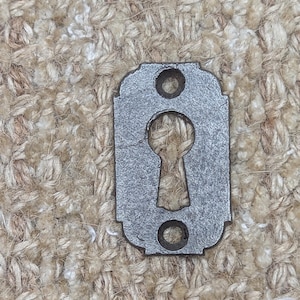 Op de afbeelding: Een zilveren sleutelgat afdekking met een decoratieve rand en twee ronde gaten. De afdekking is gemaakt van metaal en heeft een verweerde afwerking.