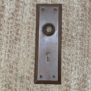 Op de afbeelding: Een rechthoekige deurplaat van verweerd brons met een centrale ronde opening voor een deurknop en een sleutelgat eronder. Het metalen oppervlak vertoont tekenen van ouderdom en oxidatie, met vier kleine schroefgaten in de hoeken.