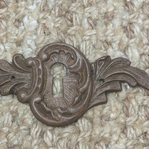 Op de afbeelding: Een decoratieve, sierlijke, bronzen sleutelgatbedekking met een bloemmotief. Het sleutelgat is zichtbaar in het midden.