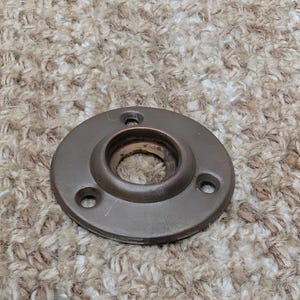 Pode incluir: Uma flange de montagem circular de metal castanho escuro com um centro elevado com uma abertura oca e três orifícios para parafusos espaçados uniformemente ao longo da borda exterior. O hardware tem um acabamento mate e liso.
