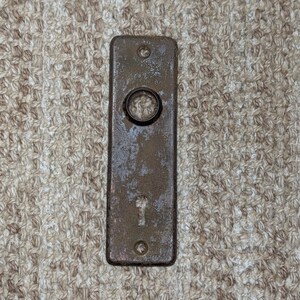 Può includere: Una placca per porta rettangolare in metallo con una finitura marrone arrugginita e usurata. La placca presenta un'apertura circolare nella parte superiore per il perno della maniglia e un foro per la chiave più piccolo sotto, con due fori.