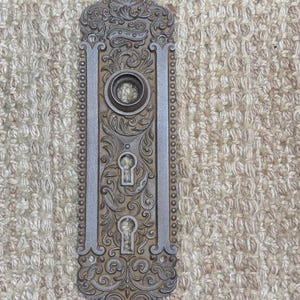 May include: Ornate, antique door plate with a detailed, symmetrical design. The metal plate features a central keyhole, surrounded by scrolling floral patterns and a decorative border. The color is a muted silver with aged patina.
