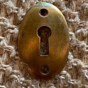 Op de afbeelding: Een ovale messing sleutelgatbedekking met twee kleine gaten aan weerszijden van het sleutelgat.