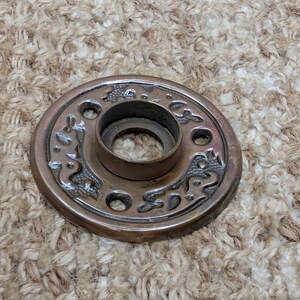 May include: A circular bronze-toned metal door plate featuring a raised central collar and three mounting holes. The surface is decorated with an embossed, swirling pattern that contrasts with the smooth, dark finish of the metal hardware.