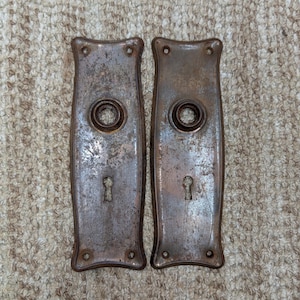 May include: Two rectangular metal door plates with a weathered, dark brown patina. Each plate features a central circular opening for a doorknob, a lower keyhole, and four small screw holes at the corners with curved edges along the sides.