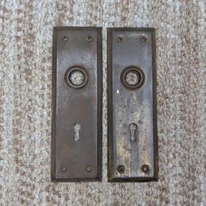 Könnte beinhalten: Zwei rechteckige Türschilder aus Metall mit einer verwitterten, bräunlich-grauen Patina. Jedes Schild verfügt über eine mittige, kreisförmige Öffnung für einen Türknauf, ein Schlüsselloch darunter und vier kleine Schraublöcher an den Ecken.