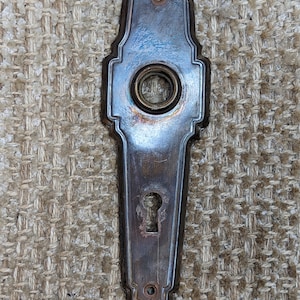 May include: Antique brass door plate with an ornate design and a keyhole. The plate has a worn, aged finish and a circular hole for a doorknob.