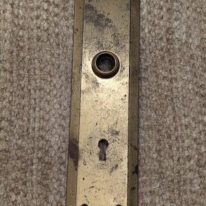 May include: A vintage brass door plate with a keyhole and a round knob. The plate has a worn, aged look and is rectangular in shape.