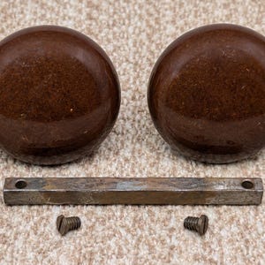May include: Two round, dark brown, glossy door knobs with a smooth finish. Included is a single rectangular metal spindle bar with a hole at each end and two small, threaded metal screws for installation, all arranged on a textured surface.