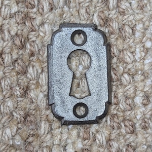 Op de afbeelding: Een zilveren sleutelgatbedekking met een decoratieve rand en twee ronde gaten. De afdekking is van metaal en heeft een licht verweerde uitstraling.