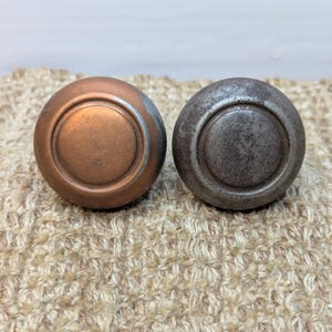 May include: Two round cabinet knobs, one is copper and the other is silver. Both have a raised ring around the center.