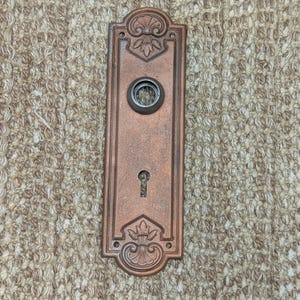 May include: A vintage-style, copper-colored door plate with an ornate design and a keyhole.