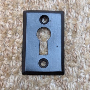 Op de afbeelding: Zwarte metalen sleutelgat afdekking met een rechthoekige vorm en afgeronde randen. De afdekking heeft twee kleine gaten aan weerszijden van het sleutelgat.