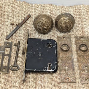 May include: Antique brass door hardware set with two ornate door knobs, a key, a strike plate, and two decorative plates. The door knobs have a cross-shaped design.