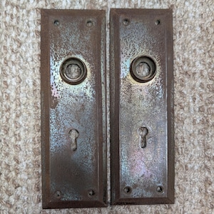 May include: Two rectangular metal door plates with a weathered, rusted finish. Each plate features a circular central opening for a doorknob and a lower keyhole cutout. The plates have four small screw holes, one at each corner, for mounting purposes.