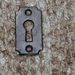 Könnte beinhalten: Eine kleine, rechteckige Metall-Schlüsselplatten-Platte im antiken Stil mit Schlüsselloch und zwei Schraubenlöchern. Die Platte hat eine dunkle, gealterte Oberfläche, was darauf hindeutet, dass sie vintage ist oder verwittert wurde. Das Schlüsselloch ist zentriert.