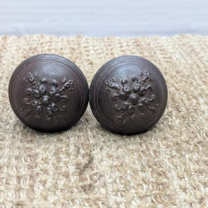 May include: Two dark brown, round cabinet knobs. Each knob features a raised floral design in the center and a ridged border. The knobs are made of a solid material and are suitable for furniture restoration or new projects.