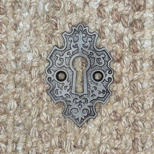 Op de afbeelding: Een zilveren sleutelgatbeslag met een sierlijk ontwerp. Het beslag bevindt zich op een beige, getextureerde achtergrond.