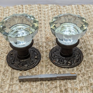 May include: Two antique glass door knobs with ornate metal bases. The knobs are clear glass with a faceted design. The bases are a dark brown metal with a floral design.