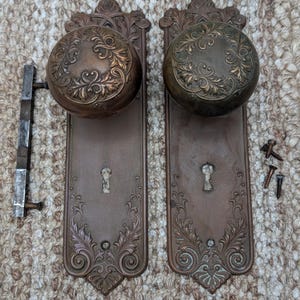 May include: Two antique bronze door knobs with ornate floral detailing. Each knob is round with a detailed pattern, mounted on rectangular plates with keyholes and decorative edges. Includes a latch and screws.